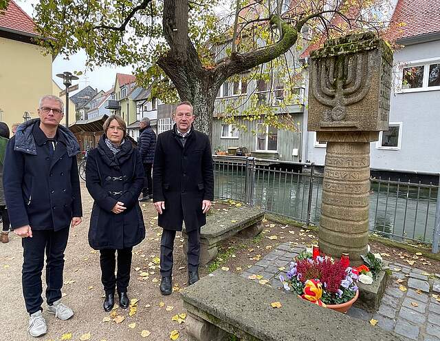 Zwei Männer und eine Frau in dinkler Kleidung stehen an einem Synagogendenkmal. Eine bunt bepflanzte Blumenschale steht am Fuße des Denkmals. Im Hintergrund befinden sich ein grpßer Baum und Häuser.