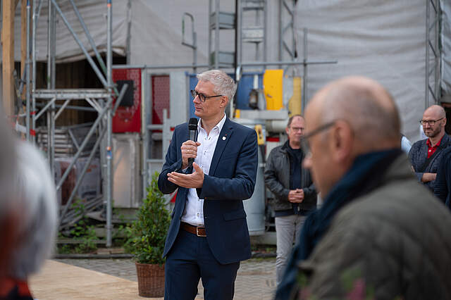 Begrüßung durch Oberbürgermeister Dr. Uwe Kirschstein vor dem historischen Rathaus zur Grundsteinverlegung. Ein Mann hält eine Rede. Im Vordergrund sieht man verschwommen Menschen. Im Hintergrund befindet sich eine Baustelle.