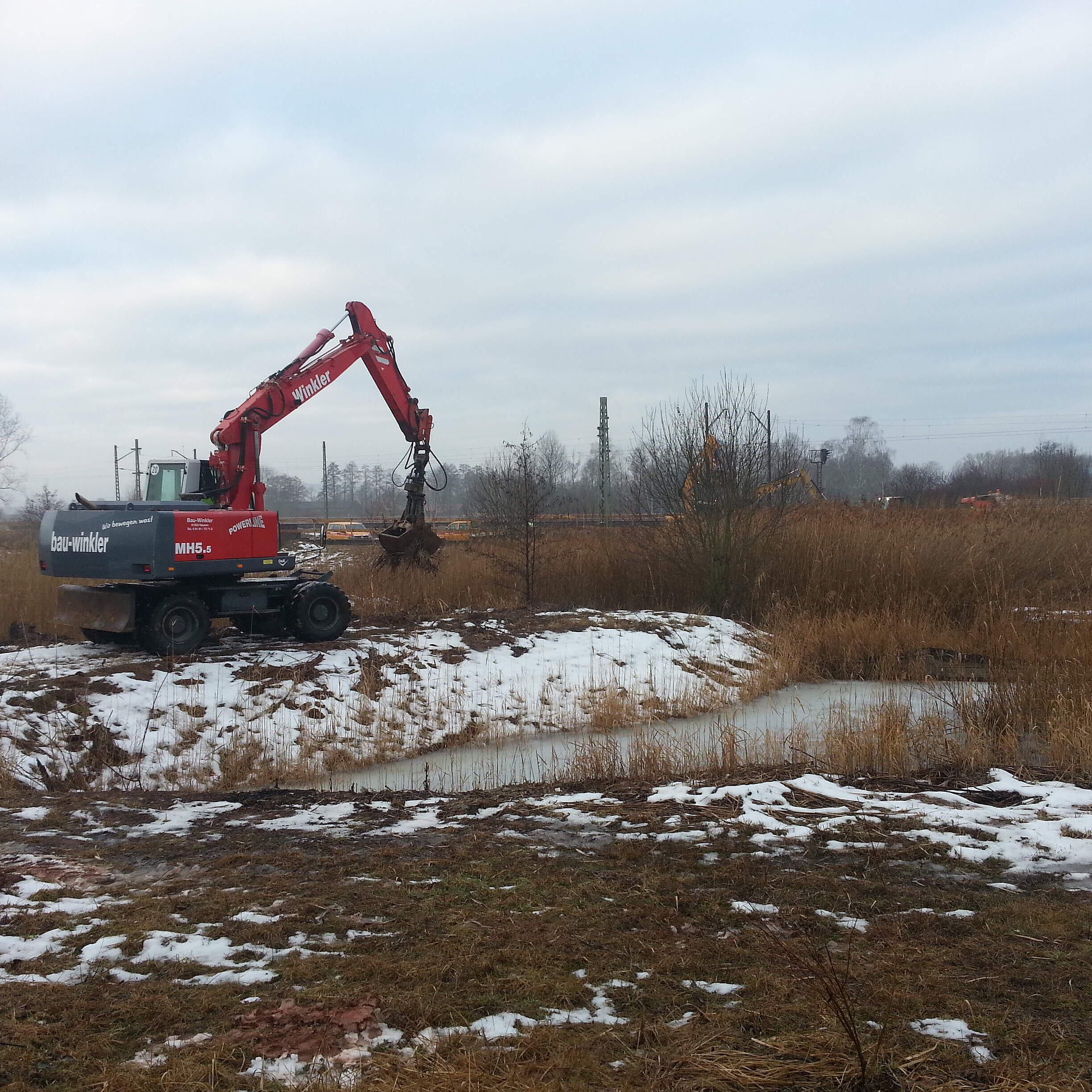 Ein Bagger gräbt in einer winterlichen Landschaft einen kleinen Teich aus.