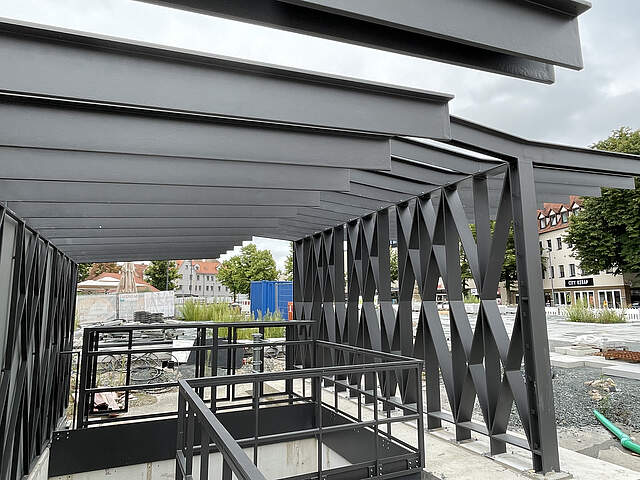 Hochbauten am Tiefgarageneingang West am Paradeplatz in Forchheim Ein moderner Hochbau aus dunklem Stahl an einem Treppenabgang zu einer Tiefgarage errichtet. Umliegend befindet sich eine Großbaustelle mit Absperrungen und verschiedenem Baumaterial.