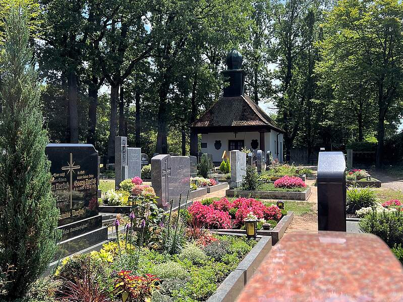 Im Hintergrund die Aussegnungskapelle am Friedhof in Kersbach. Davor bunt bepflanzte Erdgräber. Umrahmt wird das Areal von großen Bäumen.