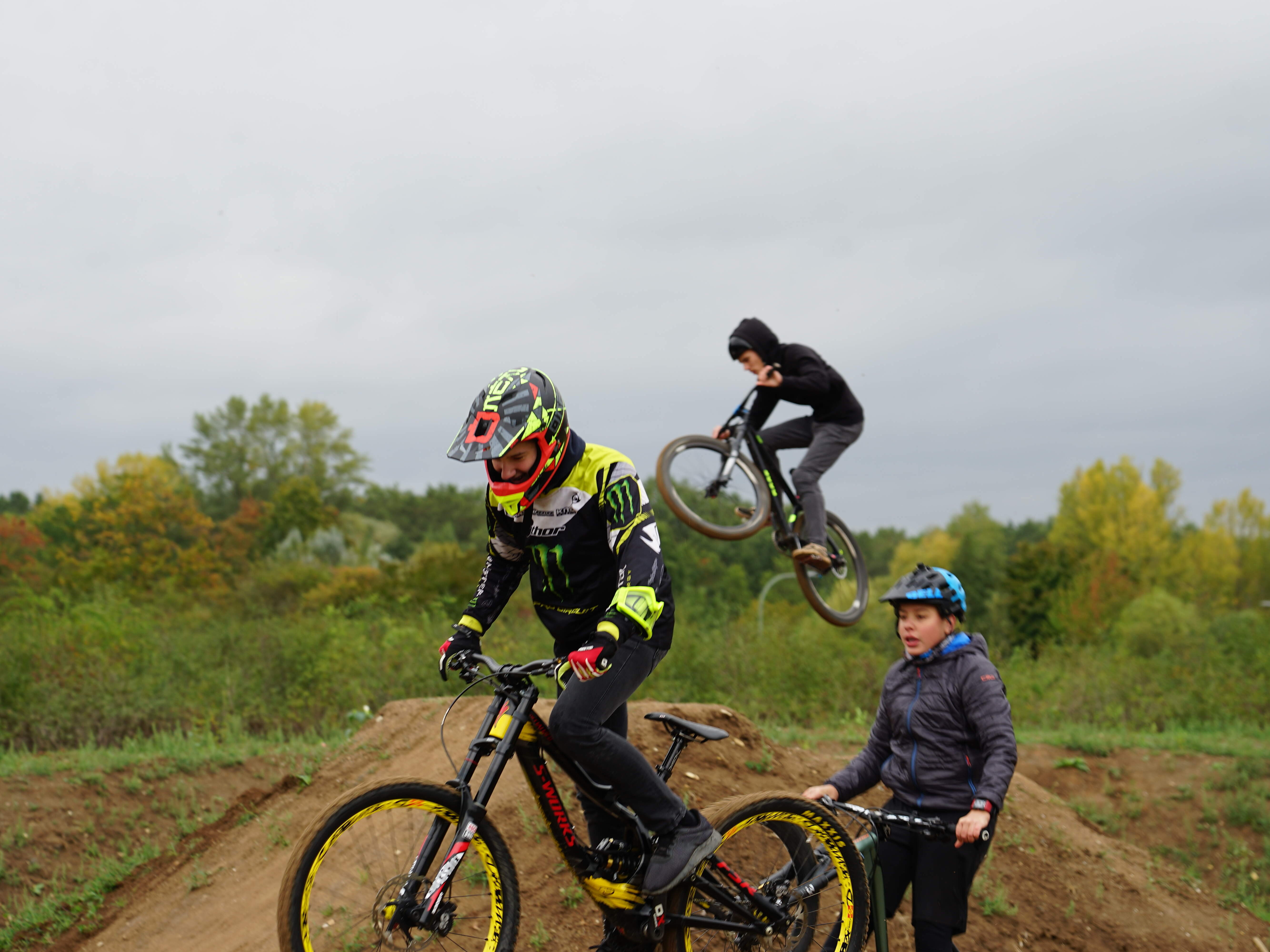 Ein Mountainbiker bei einem Sprung über einen aufgeschütteten Erdhügel im Hintergrund. Vorne sind zwei jugendliche Mountainbiker mit ihren Rädern zu sehen.