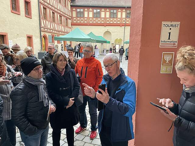 Eine Gruppe Menschen befindet sich in einem Innenhof eines großen historischen Gebäudes. Ein Mann erklärt den Gästen eine Anwendung an einem Smartphone, mit der sie sich die Stationen des neuen Jüdischen Pfades erläutern lassen können.