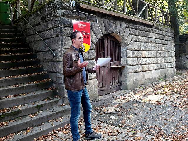 Ein Mann steht vor einer Steintreppe und Mauer vor einem Bierkeller in einem Wald. In der linken Hand hält er ein Schriftstück.