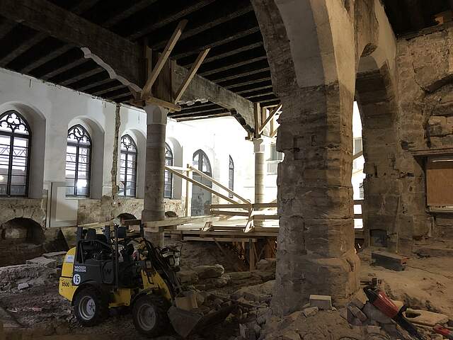 Eine Baustellen-Innenansicht in einer großen historischen Halle mit alter Holzdecke und einer Steinsäule in der Mitte der Halle. Ein kleiner Bagger und Werkzeug sind verstreut in der Halle zu sehen.