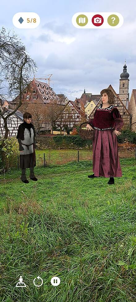 Screenshot einer Erlebnis-App, bei der zwei historische virtuelle Personen auf einer Wiese vor einer Altstadt stehen. Links im Bild eine Junge, rechts eine Frau in dunkelrotem Gewand.chnung in der linken Hand.