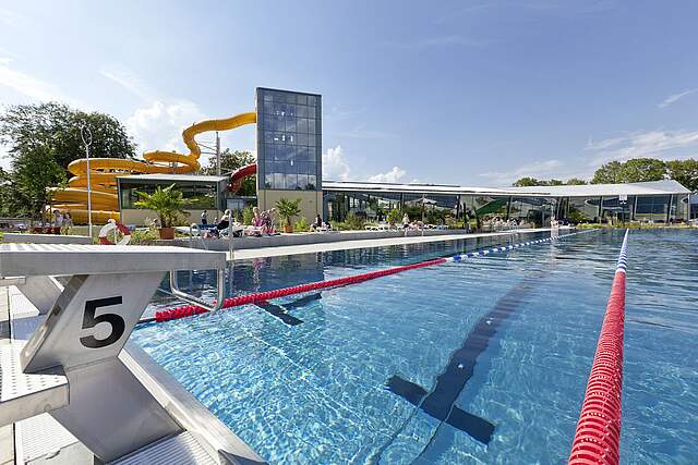 Das großer Schwimmeraußenbecken mit dem Rutschenturm im Hintergrund bei Sonnenschein.