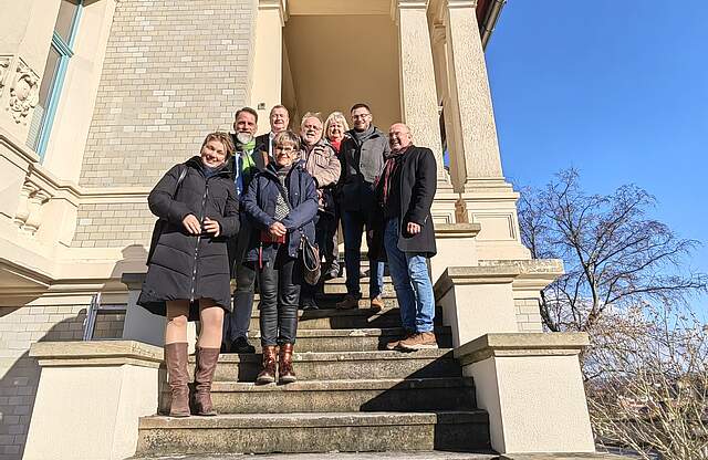 Die Delegation der Stadt Forchheim und Vertreter der Stadt Pößneck steht bei schönem Wetter auf einer Außentreppe eines großen Gebäudes.