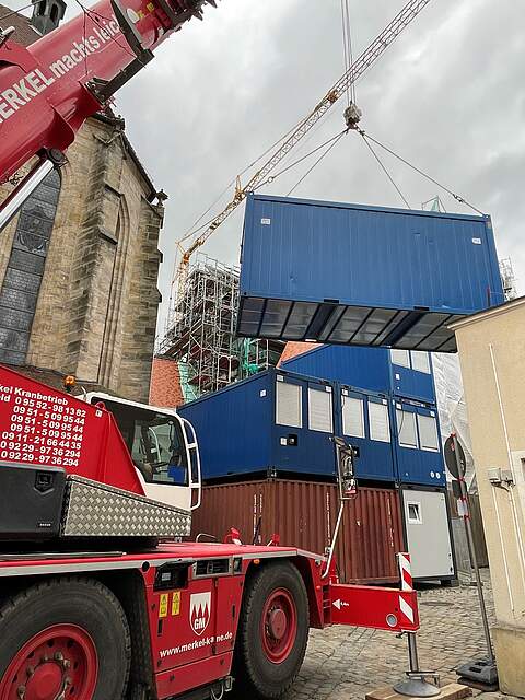 Die neuen Container für die Haustechnikgewerke an der Rathausbaustelle werden gesetzt. Ein Kran stapelt große Baucontainer übereinander.