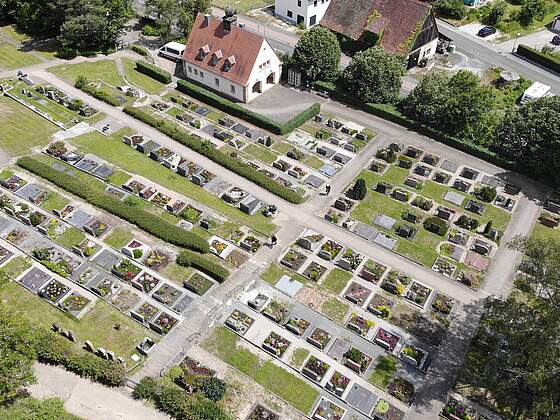 Ein terrassenförmig angelegter Friedhof mit bunt bepflanzten Erdgräbern. Treppen verbinden die einzelnen Ebenen. Links oben befindet sich eine kleine Aussegnungshalle. Im Hintergrund erkennt man Wohnhäuser.Bäume.