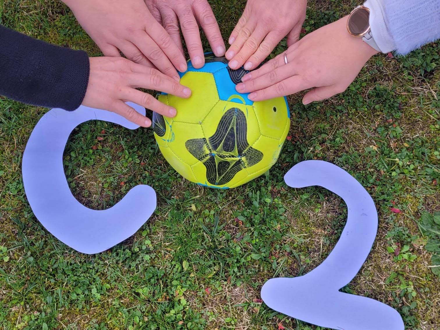 Ein ausgeschnittenes C und ein e 2 bilden zusammen mit einem Ball auf einer Wiese das Wort CO2. Fünf Hände liegen halbkreisförmig auf dem Ball.