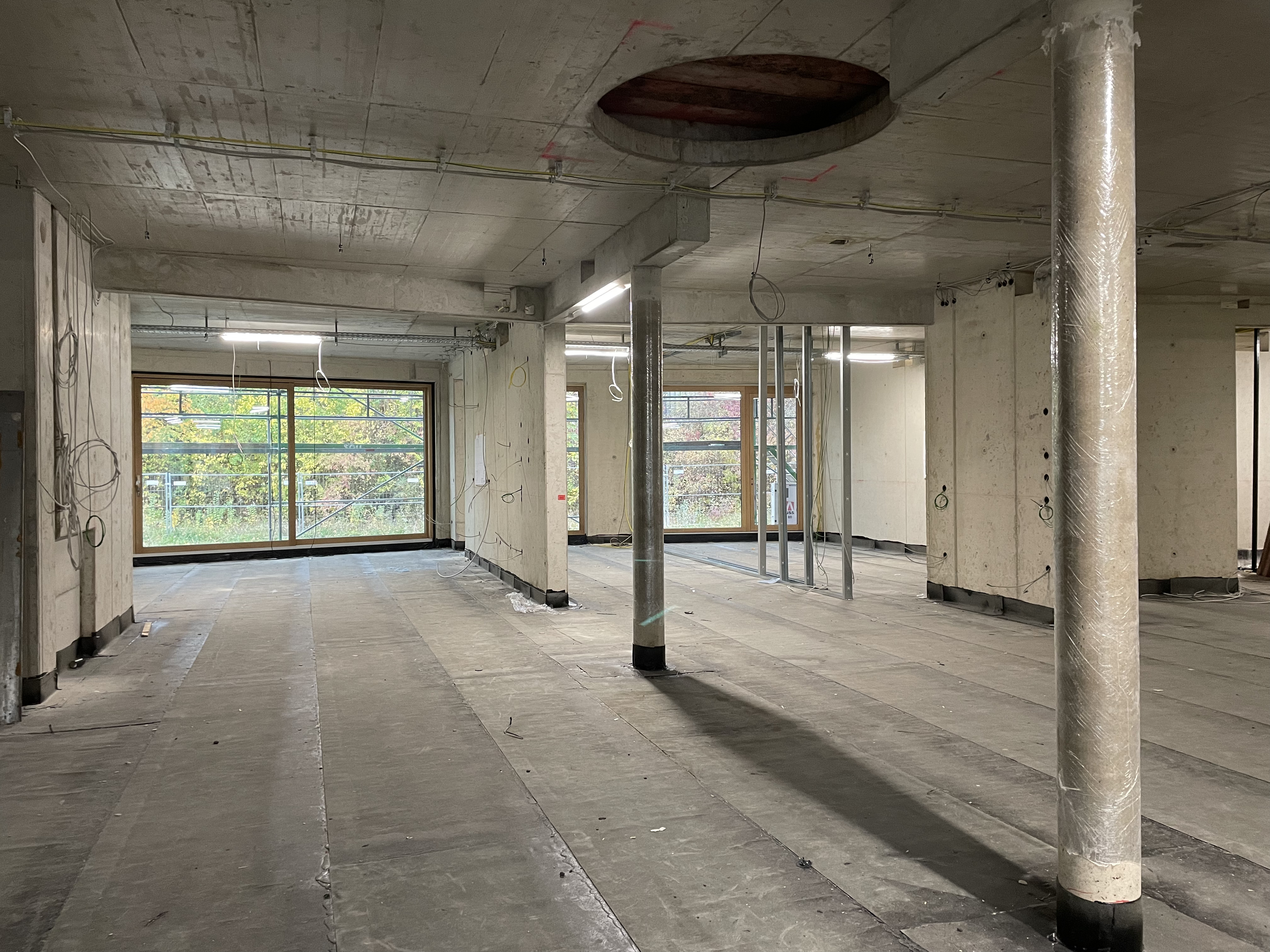 Zwei Betonsäulen und betonierte Innenwände in einem Rohbau mit großen Fensterfronten. An der Betondecke sieht man eine große runde Aussparung. Mehrere Kabel hängen von der Decke.