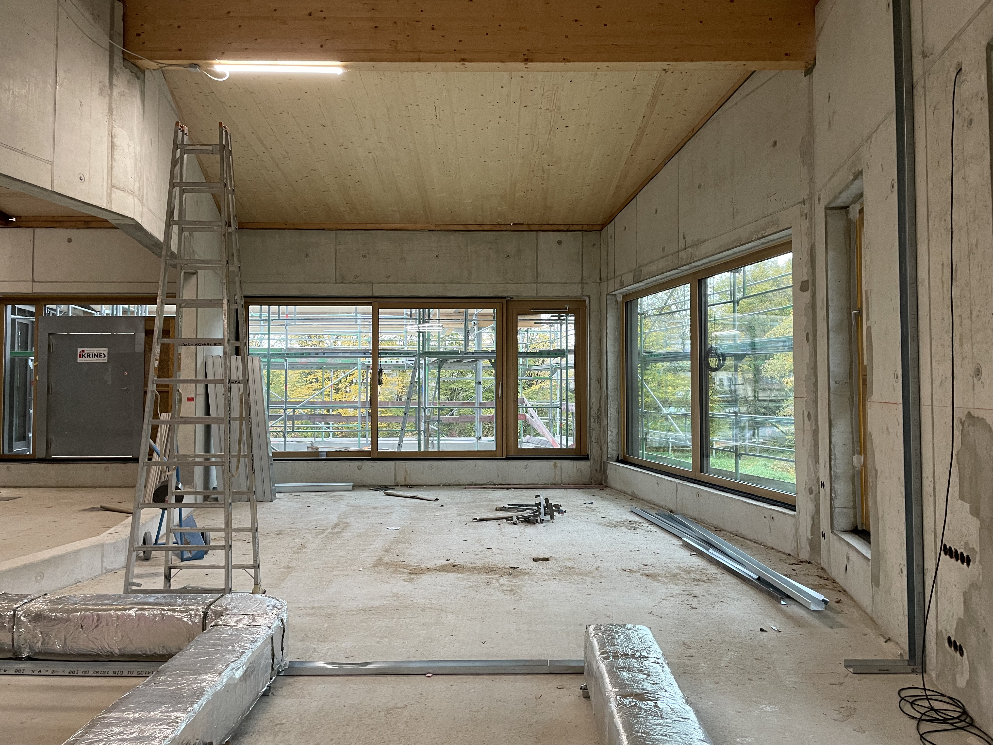 Blick von innen auf die große Fensterfront in der Raumecke in einem Rohbau. Am Boden verlaufen isolierte große Rohre. Links im Bild befindet sich eine aufgestellte Leiter unter einer Lampe.