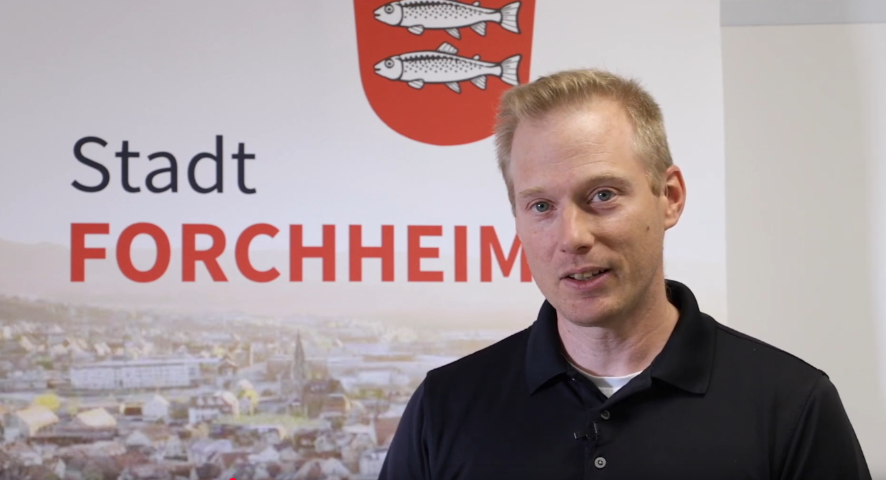 Digitaler Zwilling - Sachgebietsleiter der Geodatenverwaltung Matthias Hoffmann Sachgebietsleiter Matthias Hoffmann vor einem Plakat mit einer Luftbildaufnahme der Stadt Forchheim und dem neuen Stadtlogo.
