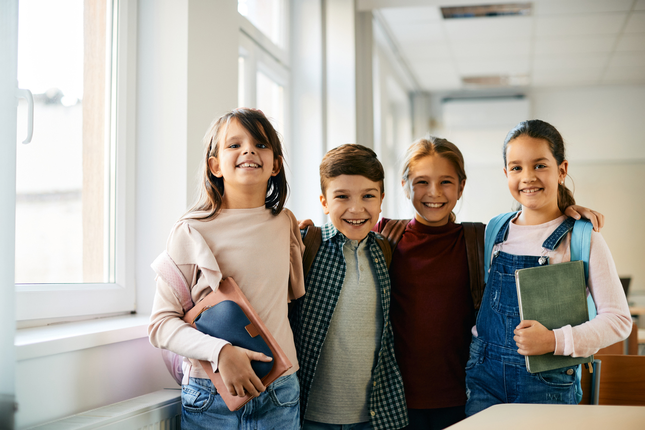 Drei Mädchen und ein Junge im Grundschulalter stehen nebeneinander in einem hellen Klassenzimmer. Das Mädchen links hält ein Schulbuch und ein Federmäppchen im Arm, das Mädchen rechts im Bild halt eine grünes Buch. Die Kinder lachen. Links im Bild befindet sich im Hintergrund eine Fensterfront.