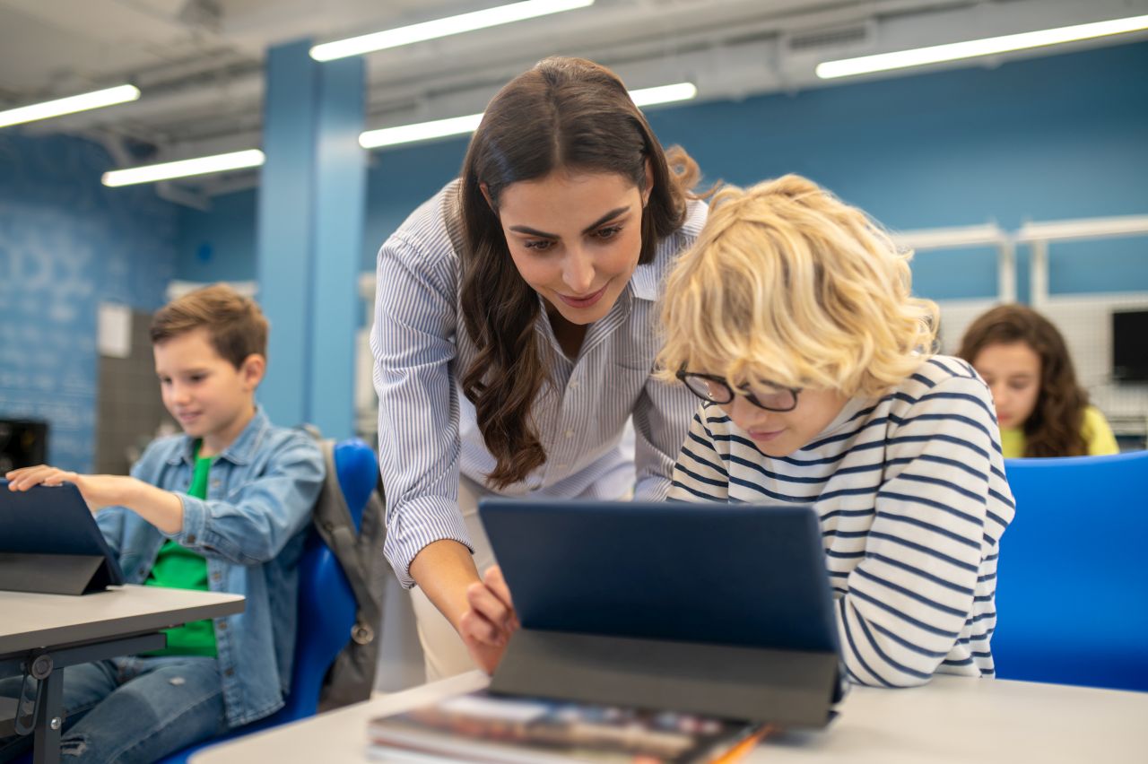 Eine Frau zeigt einem Jungen etwas auf einem Tablet. Der Junge sitzt an einem Schreibtisch in einem blau gestrichenen Raum. Links neben ihnen sitzt ein Junge mit einem Tablet an einem anderen Schreibtisch. Im Hintergrund sitzt noch ein Mädchen.