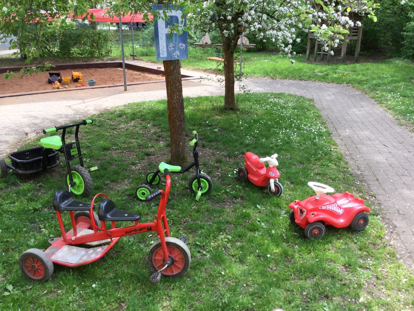 Der Kindergarten-"Fuhrpark" Mehrere Kinderräder und ein Bobbycar unter einem Baum auf eier Wiese.